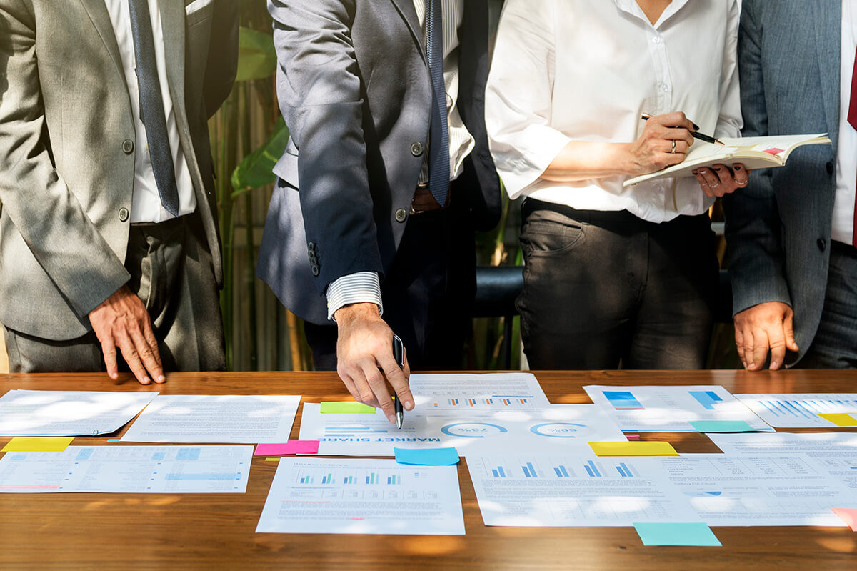 A group of business professionals standing around a wooden conference table covered in printed charts, graphs, and documents while discussing market data