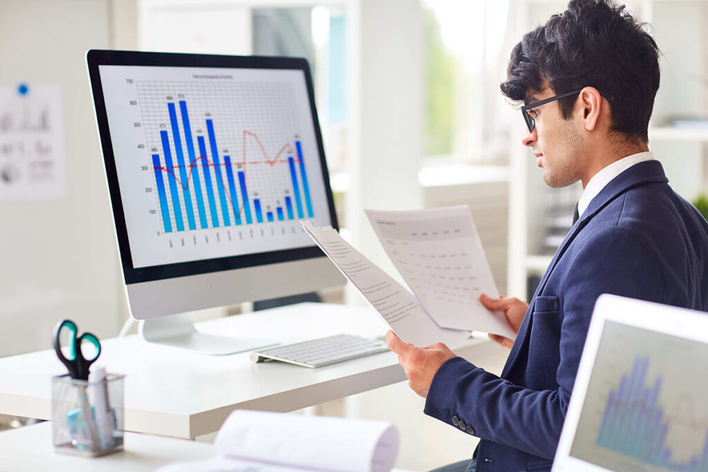A man in a suit sits at his desk reading paperwork with a desktop monitor displaying data dashboards beside him