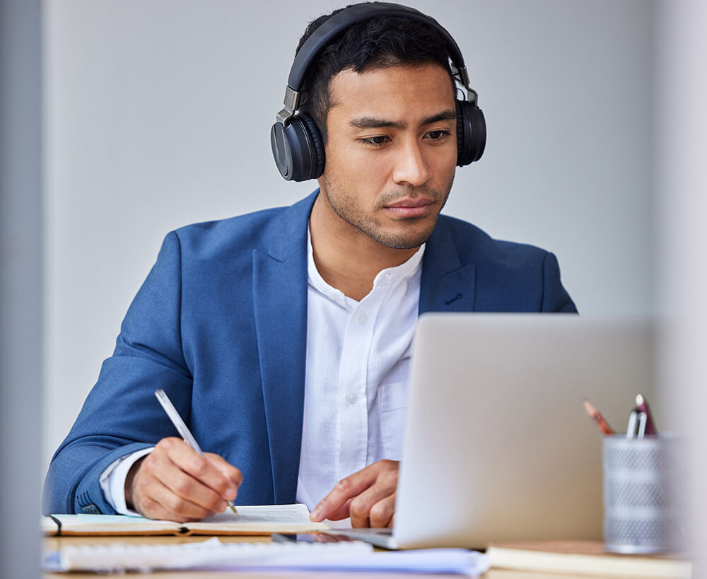 A businessman wearing black over ear headphones writing notes in a planner while focused on his laptop screen