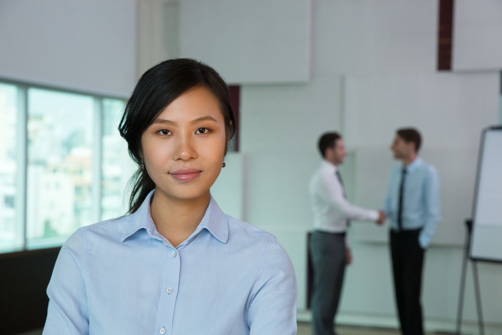 A close up of a woman in business attire, two men in business attire shaking hands in the background.