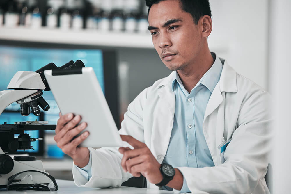A doctor in a white lab coat intently reviewing data on a white digital tablet next to a microscope in a laboratory