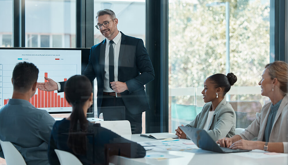 A male business executive in a suit gestures toward a digital bar chart presentation while speaking to colleagues seated around a boardroom table
