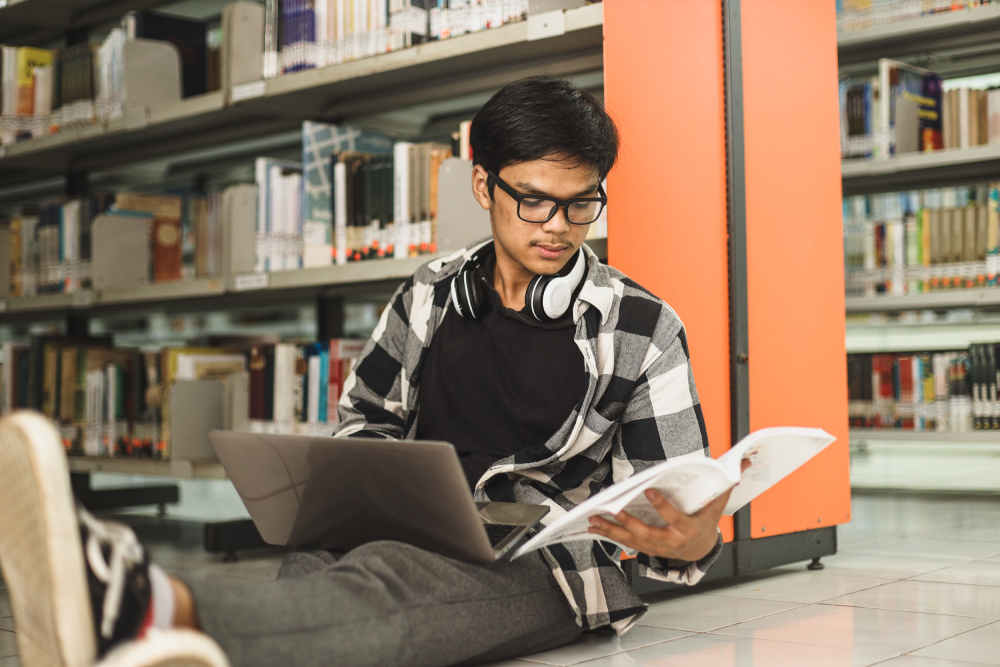 A male student studying with his laptop and a book on the floor of a library.