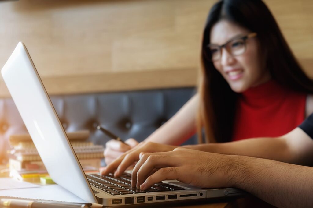 A person typing on a laptop while a smiling woman in glasses works next to them.