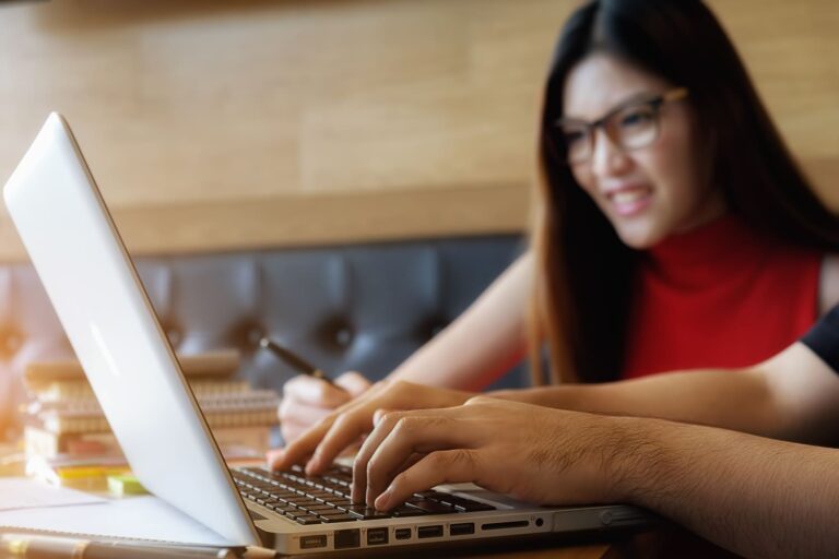 A person typing on a laptop while a smiling woman in glasses works next to them.