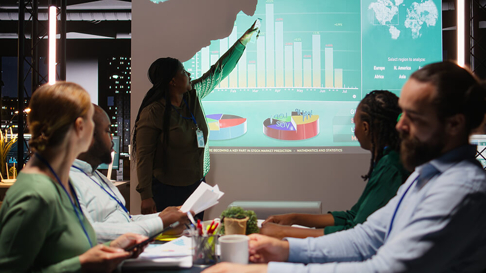 A professional pointing at a projected presentation screen with bar charts and pie graphs while presenting to a seated team in an office meeting