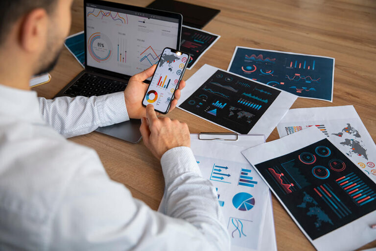 An over the shoulder view of an office worker cross referencing analytics dashboards on a smartphone, laptop, and printed reports at a wooden desk