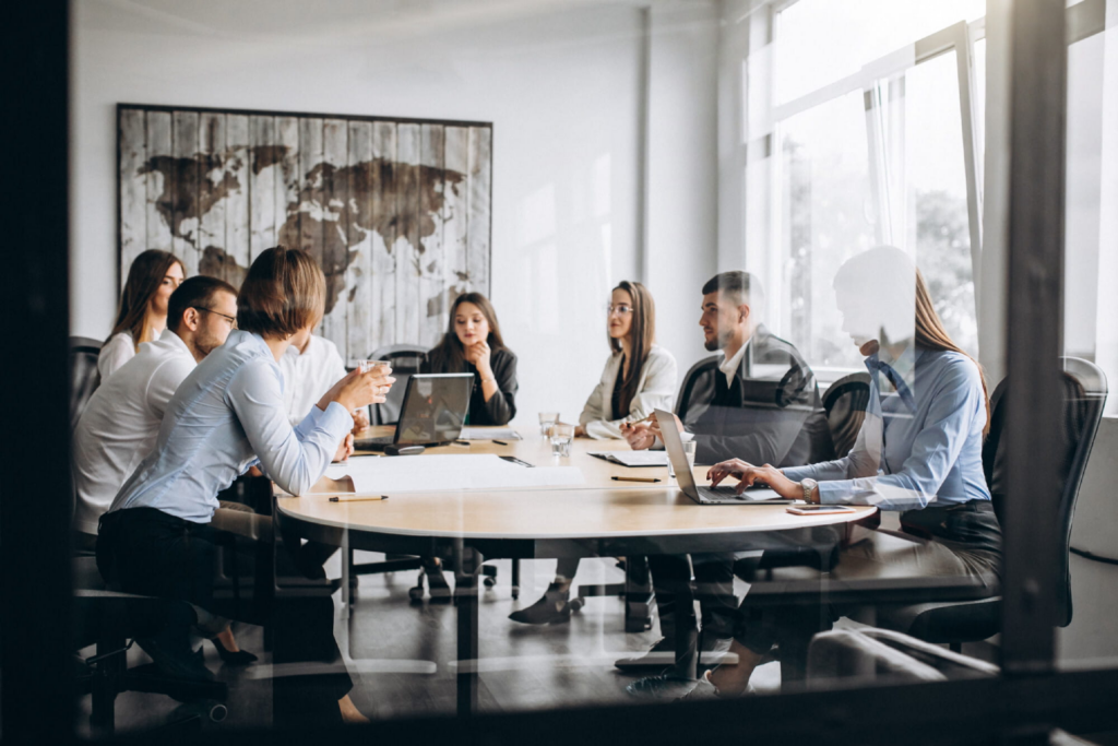 A group of corporate professionals collaborating around an oval meeting table, discussing business strategy with laptops and documents in a modern office