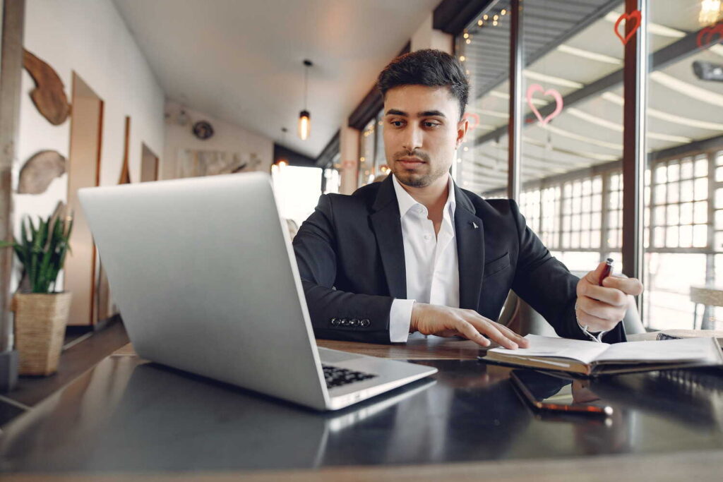Businessman in a dark suit analyzing content on a laptop while taking notes in a cafe workspace.