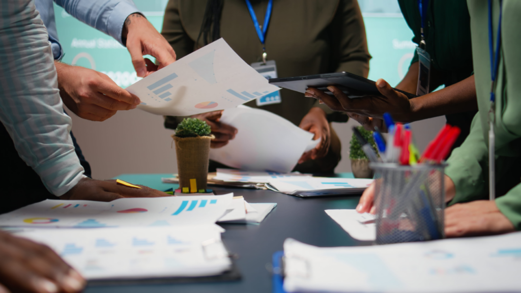 Close up of corporate team members standing around a conference table exchanging printed financial charts and digital tablet reports